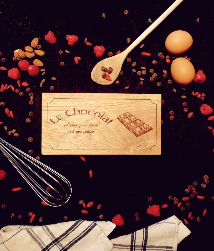 Boîte chocolat en bois massif gravée avec l'inscription 'Le Chocolat est plus qu'un plaisir, c'est une passion', fabriquée par Manufacture Dubosc et Fils. Entourée d'ingrédients pour pâtisserie : œufs, framboises, amandes et ustensiles de cuisine, idéale pour les passionnés de cuisine et les amateurs de chocolat.