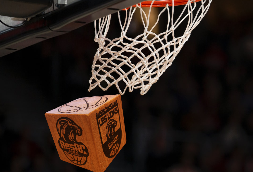 Cube en chêne massif personnalisé avec le logo du BesAC (Besançon Avenir Comtois), en train de passer à travers un panier de basket. Ce produit artisanal unique de Manufacture Dubosc et Fils est un hommage aux fans de basket et à l'artisanat français, offrant une pièce de décoration sportive authentique.