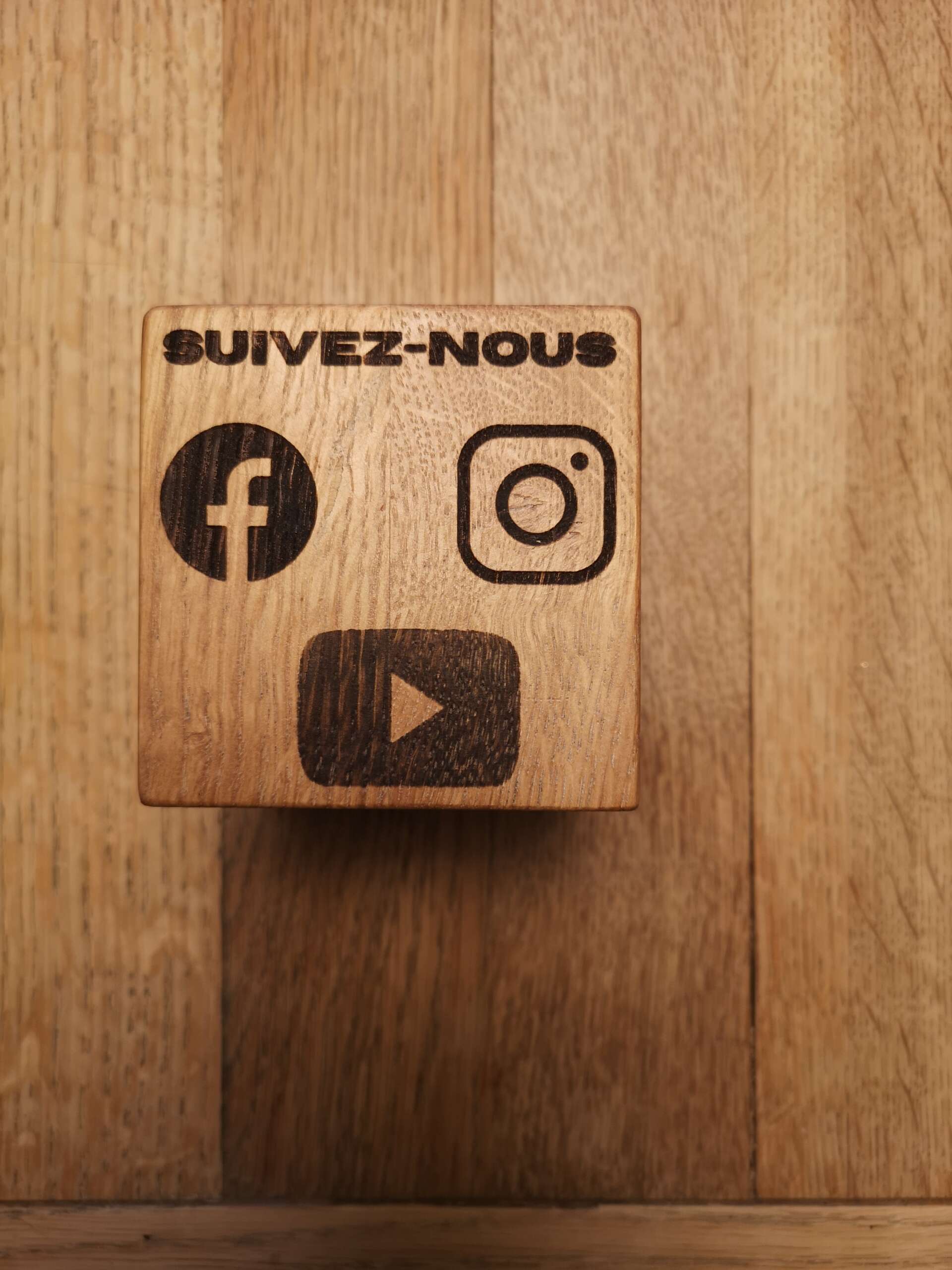 Cube en bois massif gravé avec les logos des réseaux sociaux Facebook, Instagram et YouTube, et l'inscription 'Suivez-nous'. Ce cube personnalisé est fabriqué par Manufacture Dubosc et Fils pour promouvoir la présence de l'entreprise sur les réseaux sociaux. Idéal pour une décoration moderne et informative.