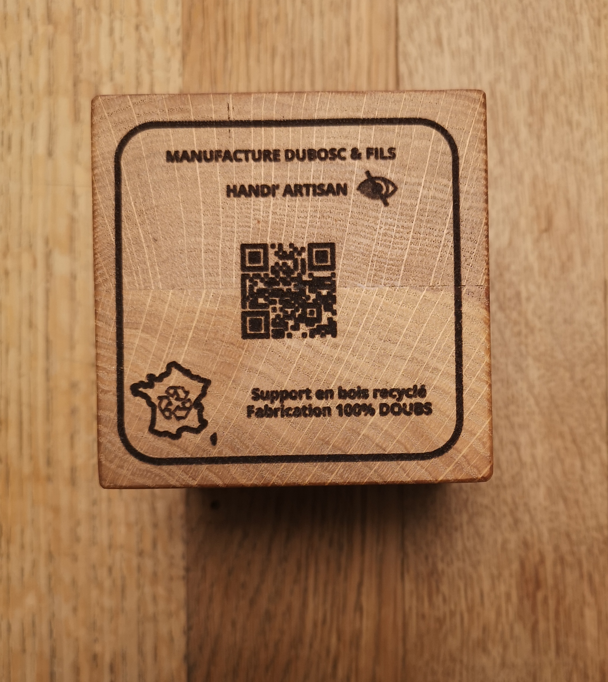 Cube en bois massif gravé avec les informations de la Manufacture Dubosc et Fils, indiquant 'Handi' Artisan' et le pictogramme malvoyant, support en bois recyclé et fabrication 100% Doubs. Présence d'un QR code pour accéder à plus d'informations sur notre entreprise artisanale et durable. Produit fabriqué à partir de bois recyclé et valorisant l'artisanat local.