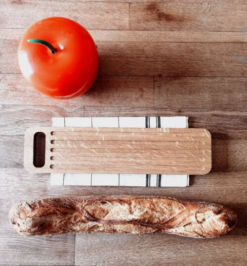 Planche à découper en chêne massif fabriquée par Manufacture Dubosc et Fils, présentée avec une baguette de pain et un récipient en forme de pomme rouge. Cette planche robuste et élégante est idéale pour trancher le pain et d'autres aliments, apportant une touche naturelle à votre cuisine tout en étant durable et pratique.