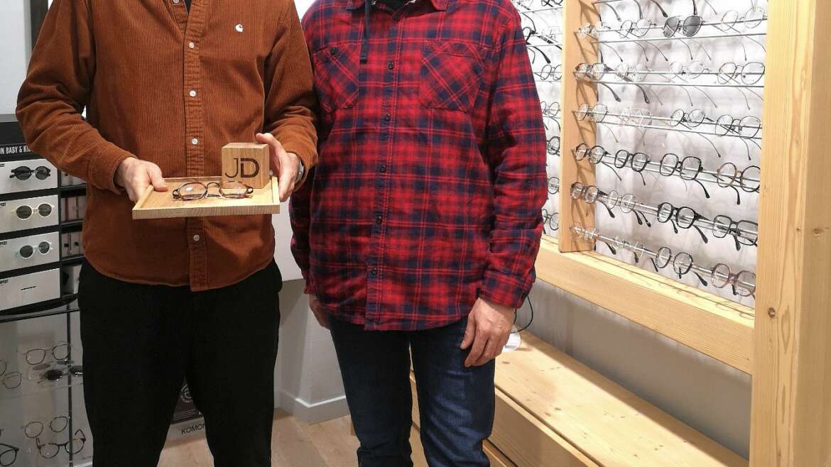 Photo de Sami Dubosc de Manufacture Dubosc et Fils avec Jimmy Dubois, opticien, tenant un plateau en chêne massif personnalisé avec un cube gravé 'JD' et une paire de lunettes. Ce produit artisanal en bois massif est conçu sur mesure, parfait pour les professionnels souhaitant un présentoir élégant et unique. Disponible sur notre boutique en ligne.