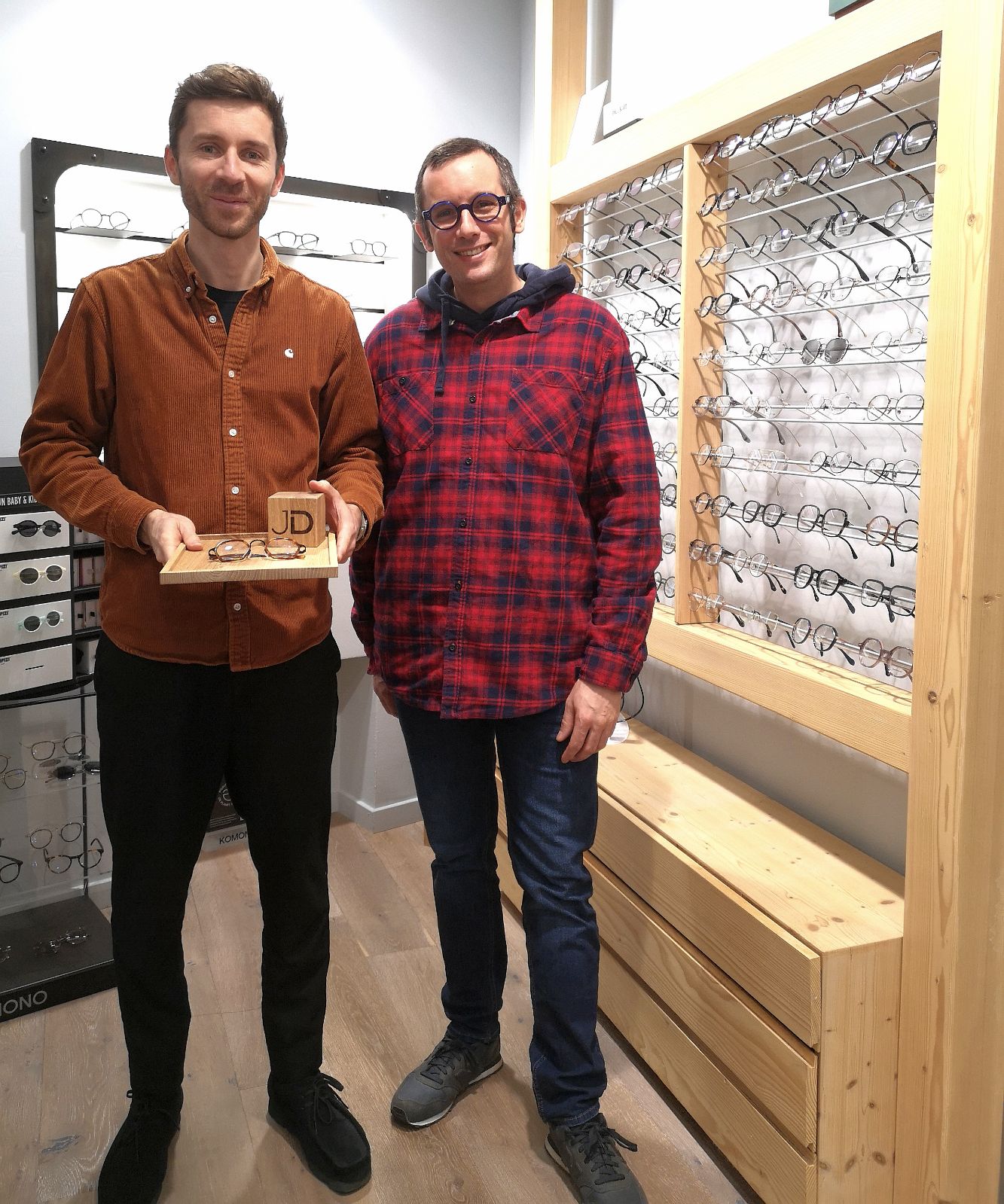 Photo de Sami Dubosc de Manufacture Dubosc et Fils avec Jimmy Dubois, opticien, tenant un plateau en chêne massif personnalisé avec un cube gravé 'JD' et une paire de lunettes. Ce produit artisanal en bois massif est conçu sur mesure, parfait pour les professionnels souhaitant un présentoir élégant et unique. Disponible sur notre boutique en ligne.