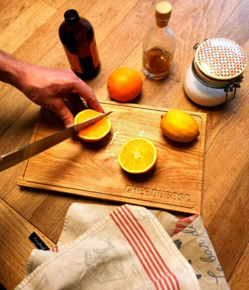 Planche à découper en bois massif personnalisée avec l'inscription 'CHEF DUBOSC', en situation d'utilisation pour couper une orange. Fabriquée artisanalement par Manufacture Dubosc et Fils, cette planche robuste est parfaite pour la préparation de fruits et autres ingrédients en cuisine. La qualité du bois massif et la personnalisation sur mesure illustrent le savoir-faire artisanal et la durabilité des produits proposés par Manufacture Dubosc et Fils.