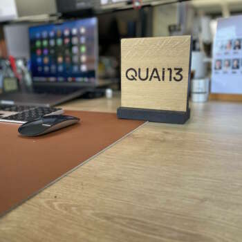 Totem en bois massif portant l'inscription 'QUAI 13', fabriqué par Manufacture Dubosc et Fils, posé sur un bureau de travail moderne. Ce totem personnalisé est un exemple parfait des créations sur mesure de notre atelier, idéales pour apporter une touche unique et professionnelle à un espace de bureau. Manufacture Dubosc et Fils propose des objets artisanaux, durables et esthétiques pour personnaliser votre environnement de travail.