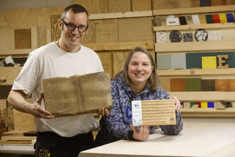 Les artisans de Manufacture Dubosc et Fils, Sami et Jennifer, posant dans l'atelier avec un morceau de bois brut et une planche à découper artisanale en bois massif, conçue et fabriquée à Deluz. Ces créations uniques mettent en valeur l'engagement de Manufacture Dubosc et Fils envers l'artisanat de qualité, la durabilité, et le savoir-faire français. Découvrez nos produits en bois personnalisés sur notre boutique en ligne et trouvez l'article parfait pour votre cuisine.