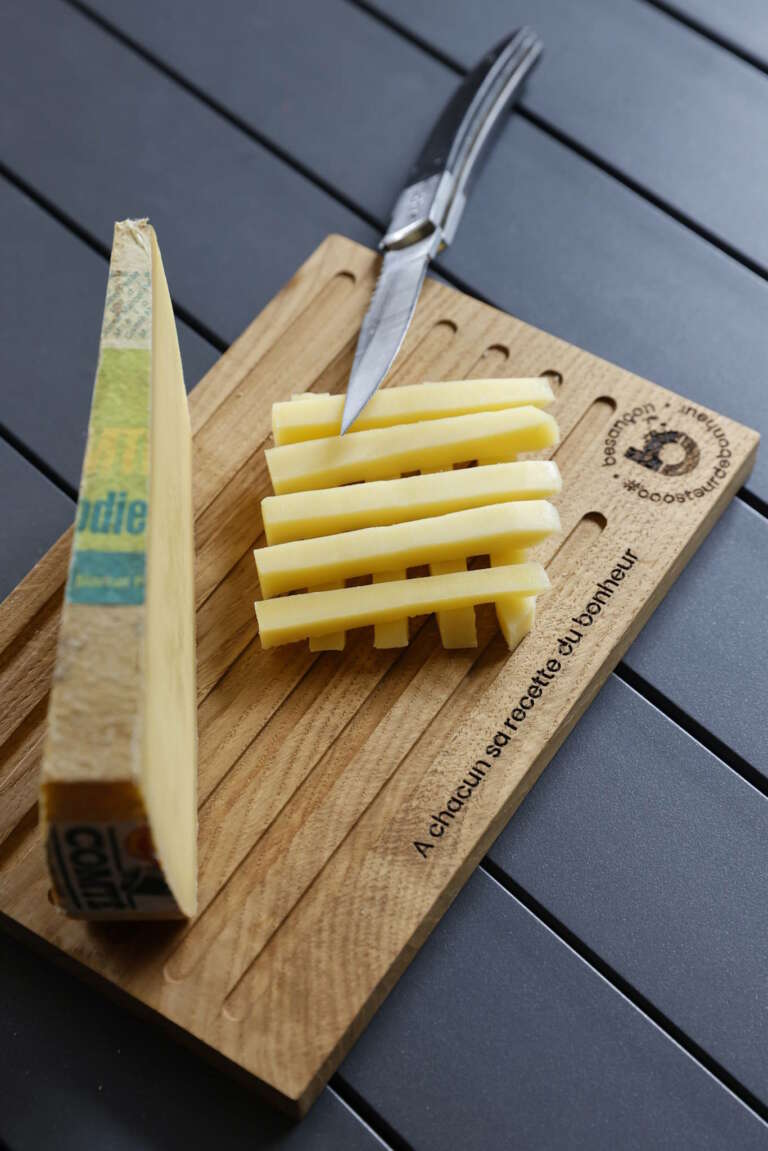 Planche à découper en bois massif de Manufacture Dubosc et Fils, utilisée pour trancher du fromage Comté avec un couteau en acier inoxydable. Ce plateau, gravé avec le message "A chacun sa recette du bonheur" et le logo "Besançon #boosteurdebonheur", allie fonctionnalité et esthétisme pour les amateurs de cuisine. Découvrez nos planches à découper artisanales et durables, parfaites pour sublimer vos moments de dégustation, disponibles sur notre boutique en ligne.