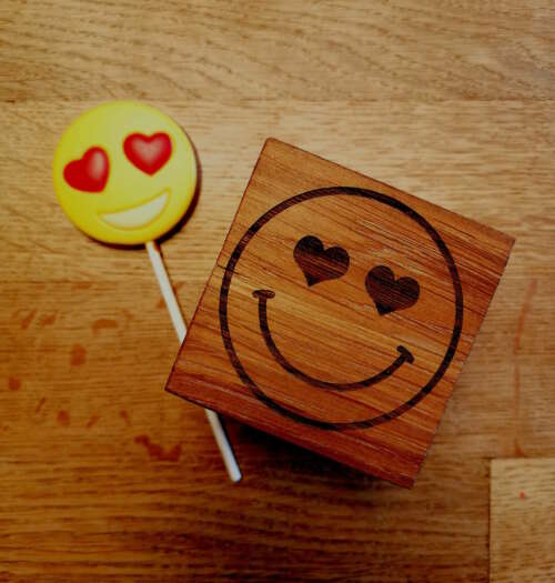 Cube en bois massif avec un emoji aux yeux en forme de cœur gravé, fabriqué par Manufacture Dubosc et Fils. À côté, une sucette emoji assortie reflète le thème de l'amour et de la joie. Ce cube est un article de décoration unique, parfait pour personnaliser un espace de travail ou une chambre, fabriqué de manière artisanale avec un souci de durabilité. Découvrez notre collection de créations en bois massif sur notre boutique en ligne.