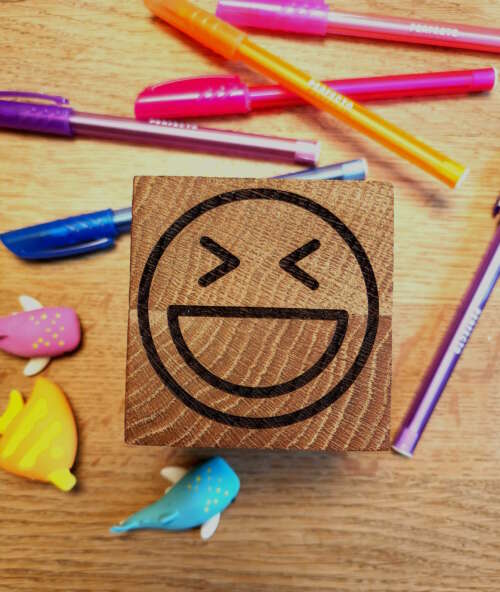 Cube en bois massif avec un emoji souriant gravé, fabriqué par Manufacture Dubosc et Fils. Entouré de stylos colorés et de petits animaux en plastique, ce cube s'inscrit parfaitement dans un univers ludique et éducatif. Réalisé avec du bois durable et gravé avec soin, il témoigne de la qualité artisanale de notre boutique en ligne. Un produit idéal pour une décoration joyeuse ou pour inspirer la créativité des enfants.