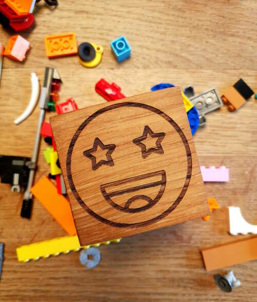 Cube en bois massif avec un emoji gravé, conçu par Manufacture Dubosc et Fils, entouré de blocs de construction colorés. Ce cube personnalisé est idéal pour apporter une touche ludique à votre décoration intérieure ou pour être utilisé comme accessoire éducatif pour enfants. Fabriqué à partir de bois durable et gravé avec précision, il reflète le savoir-faire artisanal et l'engagement de notre entreprise pour des produits éco-responsables.