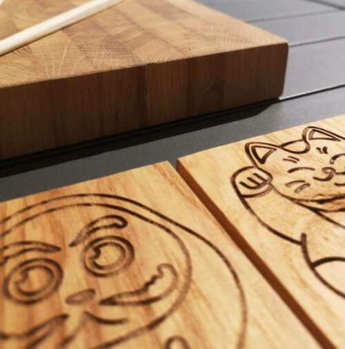 Gros plan sur une planche à découper en bois de bout de Manufacture Dubosc et Fils, accompagné de sous-verres en bois gravés avec des motifs japonais : un daruma et un maneki-neko. Ces produits, Fabrication à la main avec du bois durable , témoignent du savoir-faire artisanal de Manufacture Dubosc et Fils, et de son engagement pour des créations éco-responsables. Idéal pour apporter une touche unique et traditionnelle à votre cuisine, ces articles sont disponibles sur notre boutique en ligne pour embellir votre espace culinaire.