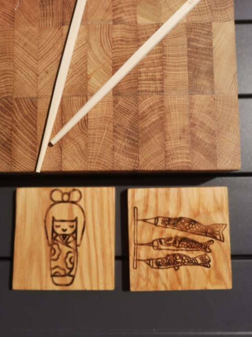 Planche à découper en bois de bout de Manufacture Dubosc et Fils avec des baguettes et deux sous-verres en bois gravés de motifs japonais : une poupée kokeshi et des poissons koï. Ces accessoires, Fabrication à la main à partir de bois durable, respecter le savoir-faire artisanal et l'engagement de notre entreprise envers des produits éco-responsables. Idéal pour une cuisine élégante et une décoration unique, disponible sur notre boutique en ligne.