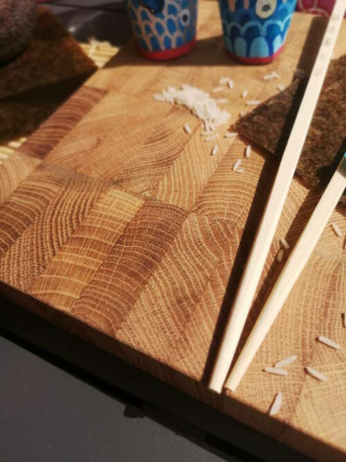 Gros plan sur une planche à découper en bois de bout de Manufacture Dubosc et Fils, avec des baguettes, des grains de riz et des feuilles de nori disposées sur la surface. Cette planche, fabriquée de manière artisanale à partir de bois durable, incarne l'engagement de Manufacture Dubosc et Fils pour des produits de qualité, faits main et éco-responsables. Parfaite pour une cuisine élégante et fonctionnelle, elle est disponible sur notre boutique en ligne pour apporter une touche unique à vos préparations culinaires.