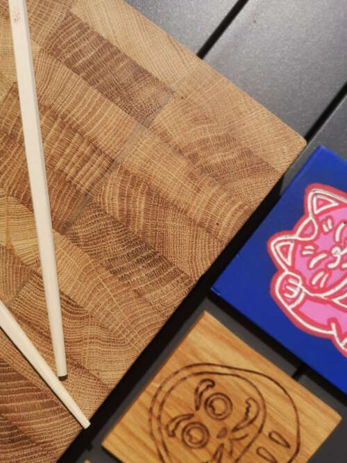 Gros plan sur une planche à découper en bois de bout fabriqué par Manufacture Dubosc et Fils, avec des baguettes, un sous-verre en bois gravé d'un motif daruma, et un dessin de maneki-neko coloré. Cette planche, conçue de De manière artisanale et à partir de bois durable, reflète le savoir-faire local et l'engagement de notre entreprise pour des produits éco-responsables. Idéale pour une décoration de table unique et élégante, cette planche est disponible sur notre boutique en ligne pour ceux. qui recherche des accessoires de qualité.