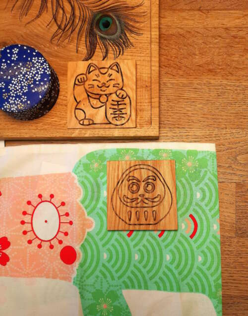 Ensemble de sous-verres en bois gravés de Manufacture Dubosc et Fils, illustrant des motifs japonais tels qu'un chat porte-bonheur maneki-neko et une figurine daruma, disposés sur une table avec un éventail et un tissu décoratif. Ces sous- Les verres sont fabriqués à la main à partir de bois durable, reflétant l'artisanat local et un engagement éco-responsable. Disponibles sur notre boutique en ligne, ces accessoires ajoutent une touche traditionnelle.