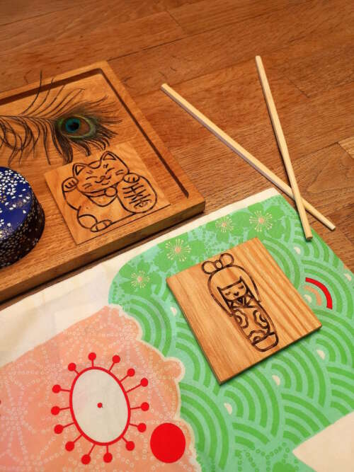 Ensemble de sous-verres en bois gravés de Manufacture Dubosc et Fils, avec des motifs japonais tels qu'un chat porte-bonheur maneki-neko et une poupée kokeshi, accompagnés de baguettes et d'un tissu décoratif. Fabriqués à la main à à partir de bois durable, ces sous-verres apportent le savoir-faire artisanal et l'engagement éco-responsable de Manufacture Dubosc et Fils. Ces produits personnalisés, disponibles sur notre boutique en ligne, ajoutent une touche unique et traditionnelle à votre décoration.