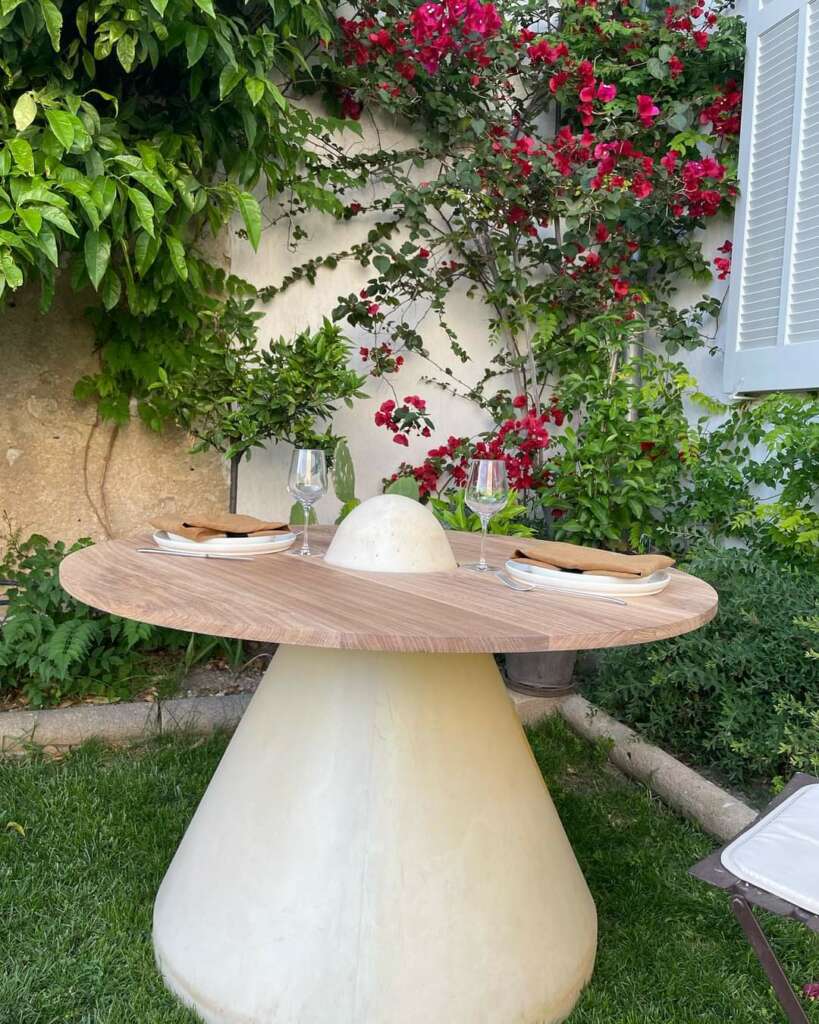 Table îlot en bois massif au design épuré, installée dans un jardin luxuriant entouré de fleurs colorées. Cette table unique est idéale pour des repas en plein air, offrant une touche naturelle et raffinée. Fabriquée de manière artisanale avec des matériaux durables, elle illustre notre engagement pour l'artisanat local et l'éco-responsabilité.