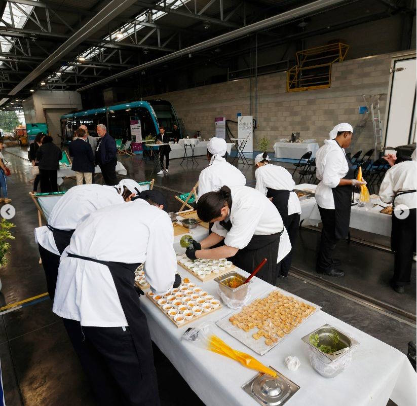 Équipe de cuisines mode d'emploi, école de Thierry Marx en train de préparer des plats lors de l'événement 'Boosteur de Bonheur' à Besançon, avec en arrière-plan un tramway et des transats pour les invités. Manufacture Dubosc et Fils soutient ce type d'événements qui mettent en avant la convivialité, le partage, et l'artisanat local. Nos produits en bois durable, conçus de manière éco-responsable et faits main, s'intègrent parfaitement dans ces moments de célébration et de qualité.