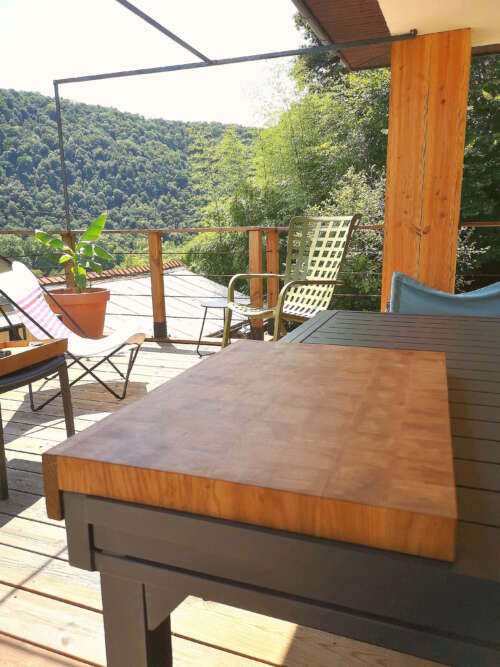 Planche à découper en bois de bout de Manufacture Dubosc et Fils posée sur une table extérieure, entourée d'une terrasse ensoleillée avec vue sur la forêt. Fabriquée de manière éco-responsable, cette planche en bois durable est faite main, issue de l 'artisanat local, et conçu avec des matériaux recyclés. Fabrication artisanale de qualité supérieure, au design intemporel, idéal pour une cuisine zéro déchet, reflétant un savoir-faire traditionnel et une conception.