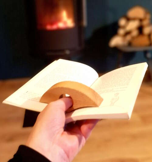 Accessoire de lecture éco-responsable en bois durable de Manufacture Dubosc et Fils, maintenant un livre ouvert devant un feu de cheminée. Fabriqué de manière artisanale, cet accessoire en bois est fait main à partir de matériaux recyclés, reflétant l'artisanat local et un savoir-faire traditionnel. Design intemporel et conception personnalisée pour une qualité supérieure, idéal pour une lecture confortable et zéro déchet.