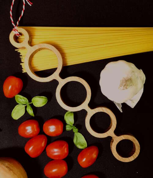 Doseur de spaghetti en bois durable de Manufacture Dubosc et Fils, entouré de tomates, basilic, ail, et spaghettis. Fabriqué de manière éco-responsable et fait main à partir de matériaux recyclés, cet accessoire de cuisine reflète l'artisanat local et un savoir-faire traditionnel. Design intemporel et fabrication artisanale de qualité supérieure, idéal pour une utilisation zéro déchet et une conception personnalisée.