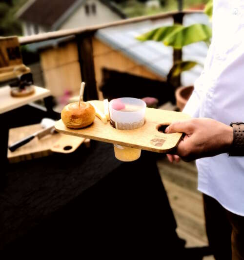 Planche apéritive en bois de Manufacture Dubosc et Fils, avec un verre de vin et des amuse-bouche, utilisée sur une terrasse en extérieur. Accessoire pratique et esthétique, parfait pour des moments conviviaux, alliant artisanat et durabilité.