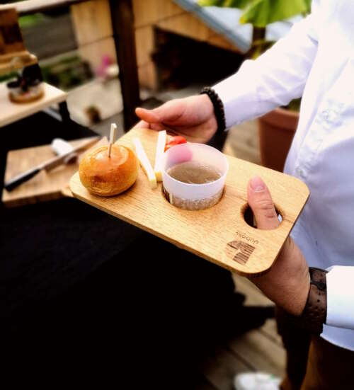 Planche apéritif en bois éco-responsable, faite main, avec un design fonctionnel permettant de tenir un verre à pied et des snacks. Accessoire en bois durable, fabriqué artisanalement à partir de matériaux recyclés, incarnant le savoir-faire local et une approche zéro déchet. Idéal pour des événements, avec une éthique de fabrication et un design sur-mesure