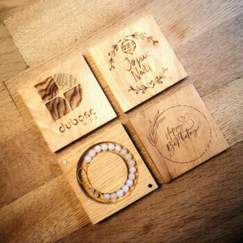 Quatre boîtes à bracelet en bois massif personnalisables, gravées avec différents motifs, dont le logo de Manufacture Dubosc et Fils, 'Joyeux Noël' et 'Happy Birthday'. Chaque boîte contient un bracelet en pierre de lune, parfait pour des cadeaux uniques et Ces créations artisanales en bois sont idéales pour les événements spéciaux ou les cadeaux d'entreprise, reflétant l'engagement de notre boutique en ligne dans la qualité et la durabilité.