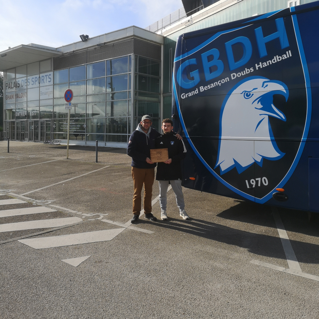 Étienne et Sami Dubosc, représentants de la Manufacture Dubosc et Fils, posent devant le bus officiel du Grand Besançon Doubs Handball (GBDH) avec un plateau en bois massif gravé, symbole d’engagement local et artisanal. Ce plateau, conçu pour récompenser l’homme du match, illustre le savoir-faire durable et l’excellence artisanale de la Manufacture.