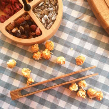 Pince en bois massif de Manufacture Dubosc et Fils, posée sur une nappe à carreaux avec des popcorns et un plateau compartimenté de fruits secs et graines. Cet accessoire en bois naturel est élégant et pratique, parfait pour servir des aliments avec raffinement.