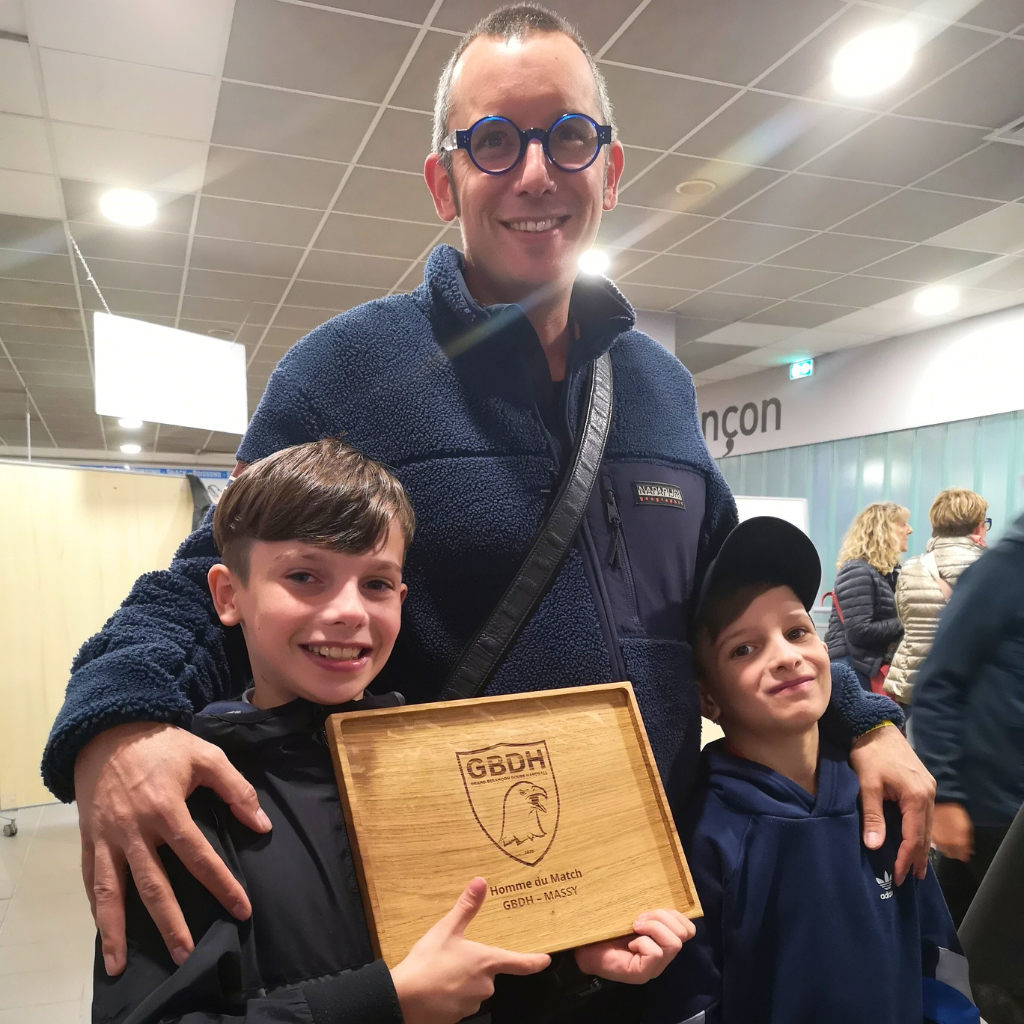 Moment de transmission entre générations au Grand Besançon Doubs Handball (GBDH) : un artisan de la Manufacture Dubosc et Fils pose avec ses enfants, tenant fièrement le plateau en chêne massif gravé récompensant l’homme du match. Une création unique qui incarne les valeurs du sport, du savoir-faire artisanal et de la durabilité.