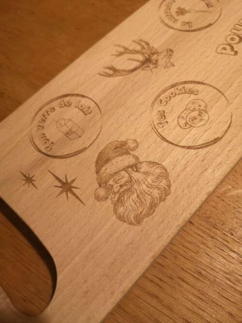 Détail d'une planche de Noël en bois massif gravée par Manufacture Dubosc et Fils, montrant des illustrations de Père Noël, d'un renne, et des emplacements dédiés aux biscuits et au lait pour le Père Noël. Cette création artisanale est conçue pour ajouter une touche de magie et de tradition à vos fêtes de fin d'année. Un cadeau parfait, fait main et durable, qui allie qualité et esprit de Noël.