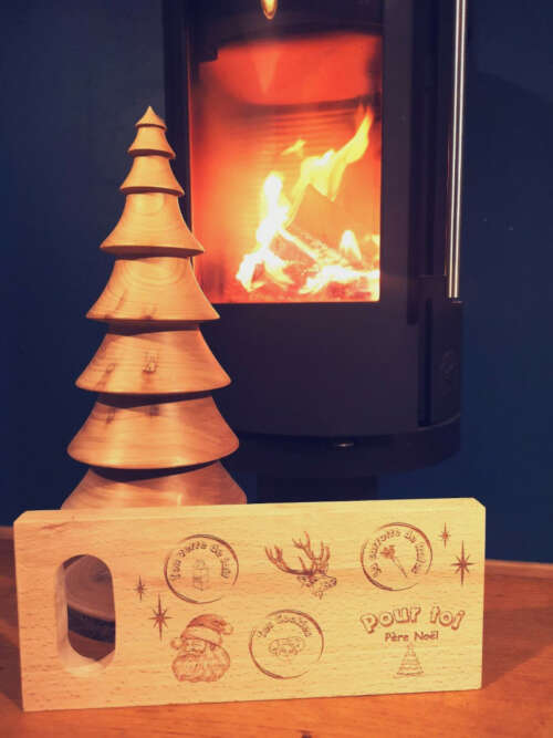 Planche de Noël en bois massif gravée, accompagnée d'un sapin en bois, devant un feu de cheminée créant une ambiance chaleureuse et festive. Créée par Manufacture Dubosc et Fils, cette planche personnalisée est parfaite pour célébrer les fêtes avec authenticité et charme artisanal. Un cadeau unique fait main, idéal pour décorer votre intérieur et apporter une touche de magie à Noël.