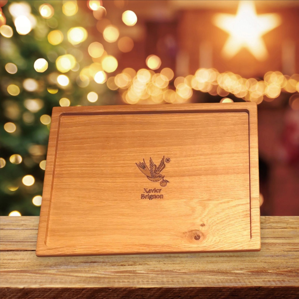 Plateau en bois massif gravée, conçue artisanalement par Manufacture Dubosc et Fils, présentée dans une ambiance festive avec un arrière-plan illuminé par des guirlandes et un sapin de Noël. Ce produit unique, personnalisé avec le logo du maître chocolatier Xavier Brignon, est parfait pour les fêtes de fin d'année. Alliant artisanat local et design élégant, cette planche est idéale pour sublimer vos présentations culinaires ou offrir un cadeau personnalisé inoubliable. Découvrez nos créations en bois sur-mesure sur notre boutique en ligne.