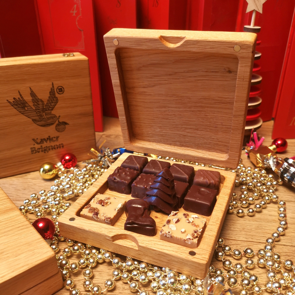 Une élégante boîte en chêne massif personnalisée , conçue par Manufacture Dubosc et Fils , contenant de délicieux chocolats artisanaux réalisés par le maître chocolatier Xavier Brignon. La boîte gravée en bois local est mise en valeur dans un décor festif avec guirlandes dorées et chocolats soigneusement disposés. Cette création unique célèbre l'artisanat d'exception et le savoir-faire régional, idéale pour les cadeaux d'entreprise ou les fêtes de fin d'année. Découvrez nos boîtes en bois massif personnalisées et cadeaux artisanaux sur notre boutique en ligne.