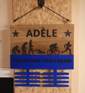 Gros plan sur un présentoir de médailles en bois massif personnalisable, gravé avec le prénom "Adèle" et des illustrations représentant l'évolution du triathlon : natation, cyclisme, et course à pied. Le support est complété par une barre bleue indiquant "Evolution Triathlon" et des crochets en bois pour suspendre les médailles. Fabriqué avec soin par la Manufacture Dubosc et Fils, ce produit allie design élégant et fonctionnalité, parfait pour les sportifs passionnés.