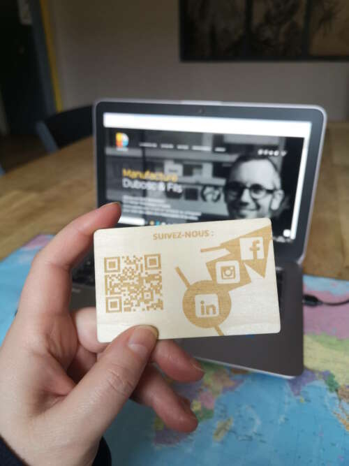 Carte de visite en bois massif personnalisée avec gravure laser, incluant un QR code renvoyant sur le site de la Manufacture Dubosc et Fils. En arrière-plan, l'ordinateur affiche le site officiel de cette entreprise artisanale basée à Deluz, spécialisée dans la création d'accessoires en bois écoresponsables et sur-mesure. Un exemple concret d'éco responsabilité et d'originalité au service de la communication professionnelle.