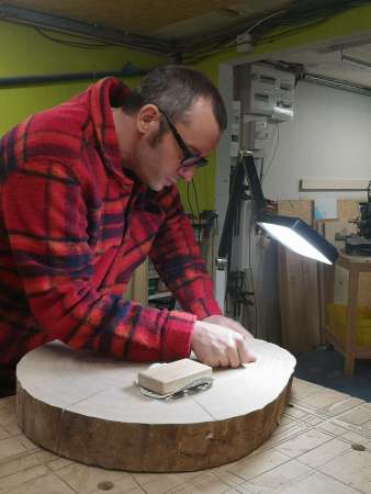 Sami Dubosc Artisan de Manufacture Dubosc et Fils travaillant minutieusement sur une tranche de bois massif pour la création d'une table rustique. Le soin apporté au ponçage et aux finitions reflète l'engagement de notre atelier pour la qualité artisanale et la personnalisation de chaque pièce en bois. Découvrez nos créations uniques sur notre boutique en ligne pour apporter une touche naturelle et durable à votre intérieur.